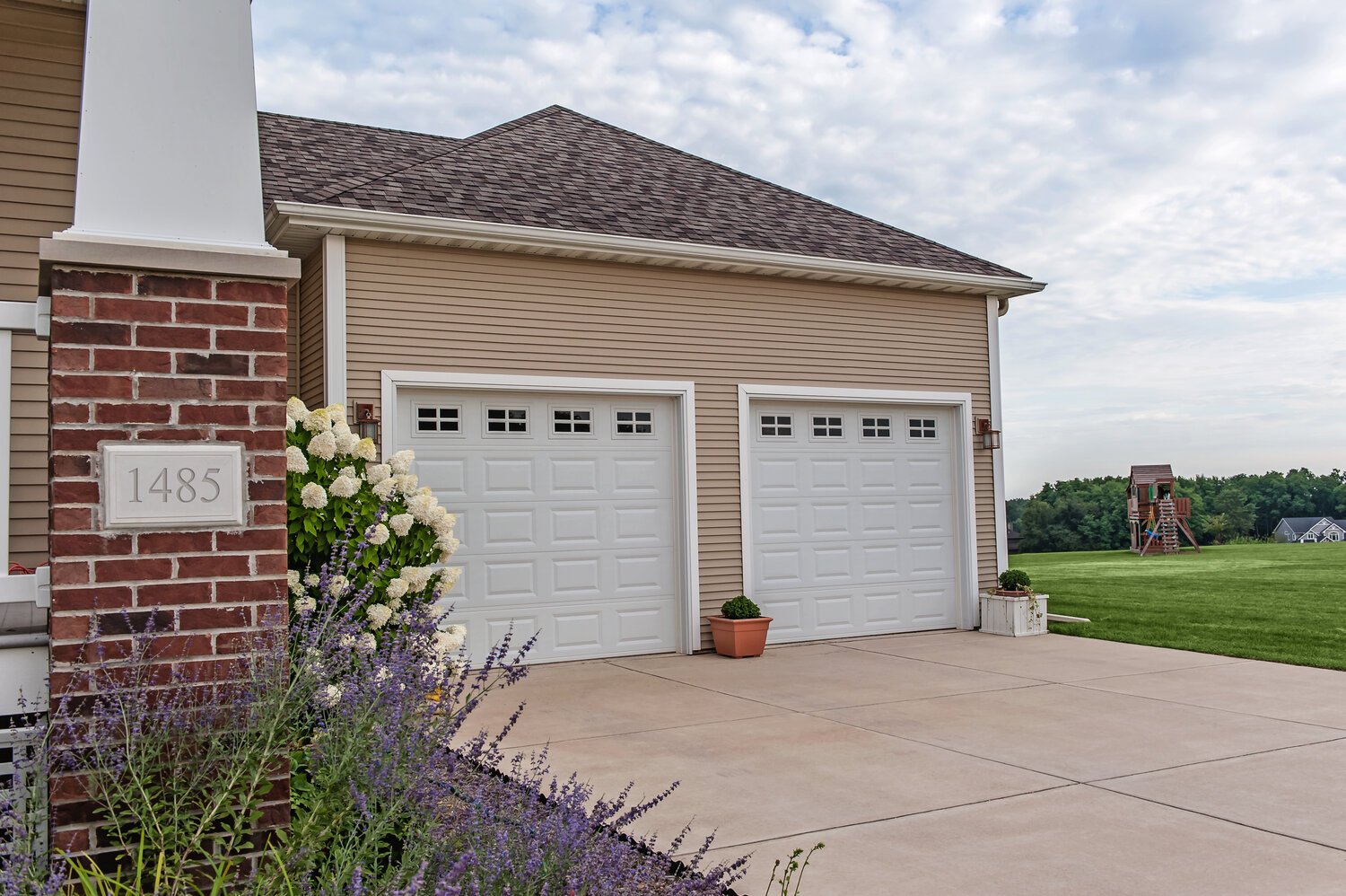 Two-car garage with beige siding, brick pillar, and a flower bed with purple blooms in the foreground.